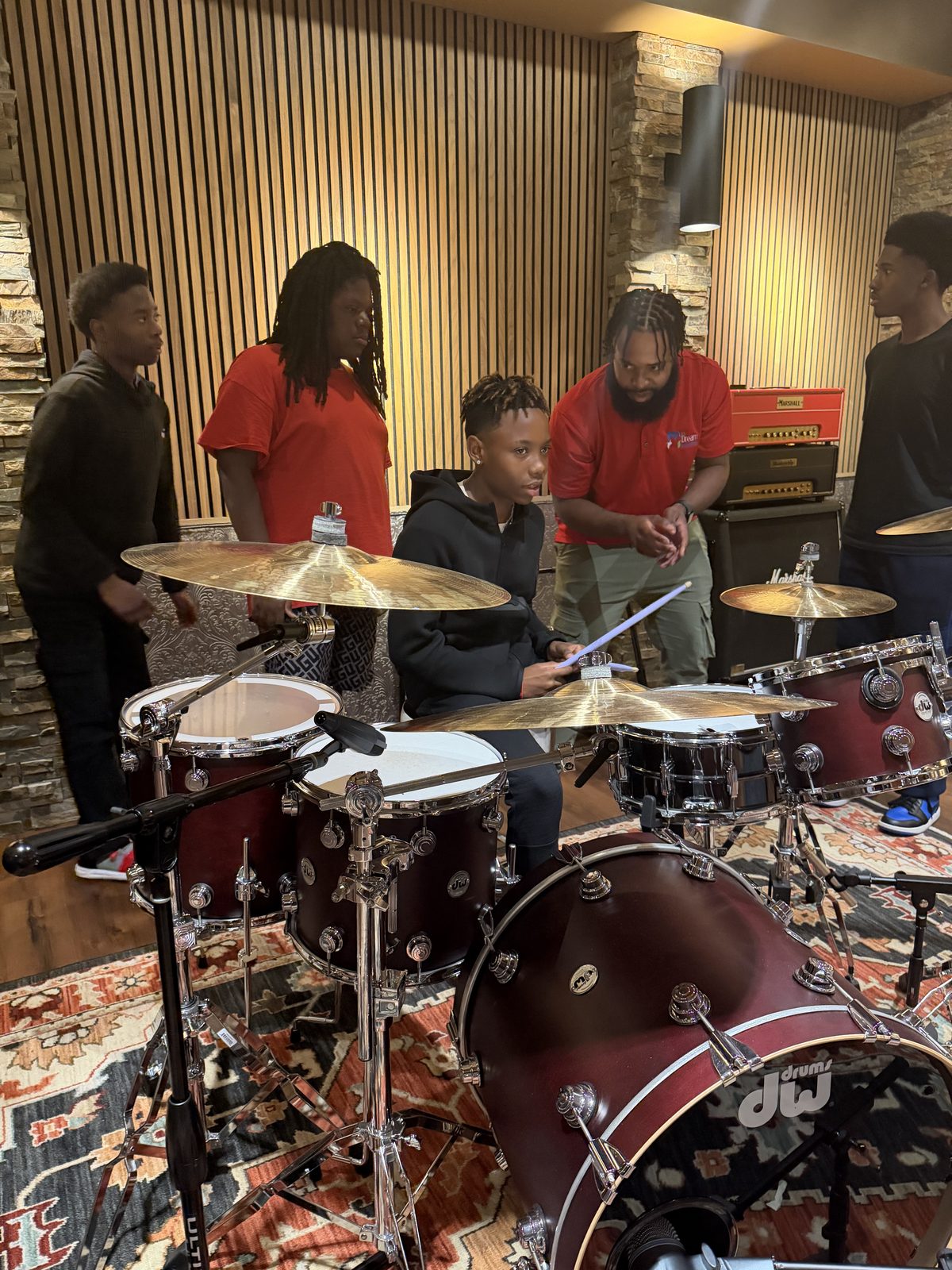 Student playing a DW drum kit in a professional studio during a Kinetic Vibez session — instructor guiding, peers watching