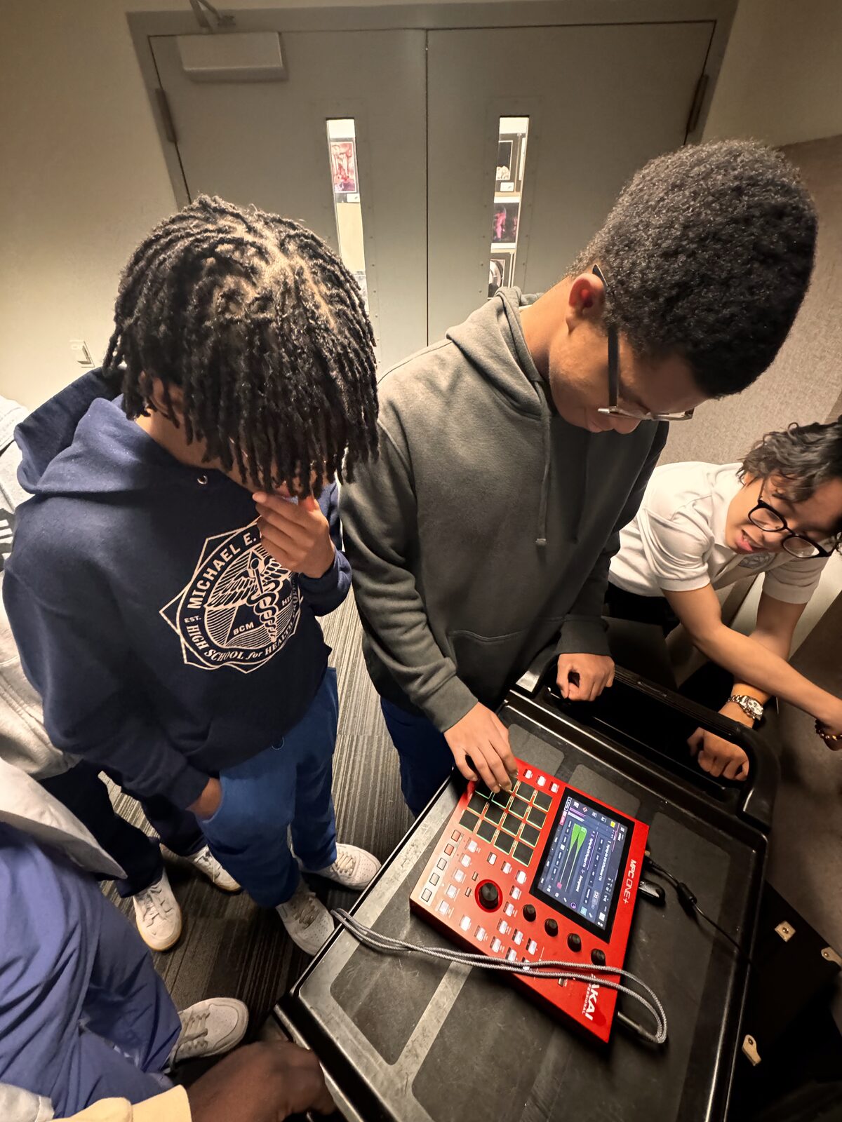 Three students crowded around an Akai MPC beat machine, hands-on during a Kinetic Vibez music production residency