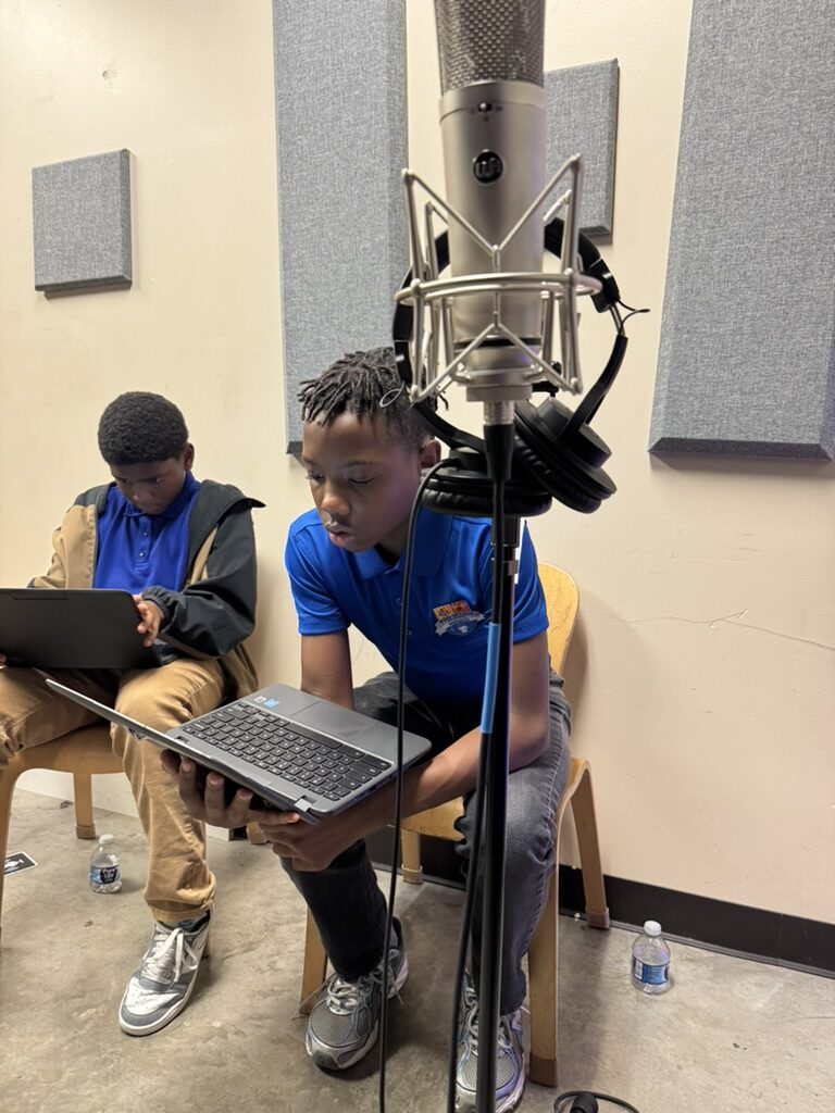 Students in school uniforms recording at a studio microphone, referencing lyrics on a laptop during a Kinetic Vibez songwriting session